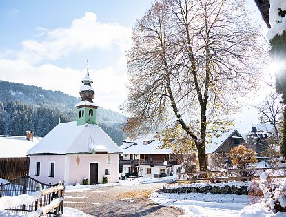 Verschneite Ortschaft Kelchsau im Winter, Kapelle und kahler großer Baum bei Sonnenstrahlen